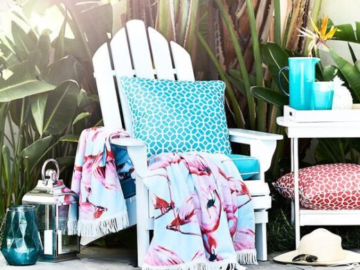 Vibrant flamingo towel on white Adirondack chair by sunny pool with tropical plants.